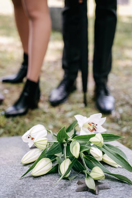 A close-up of a floral arrangement featuring white lilies with open blooms and unopened buds, complemented by long, slender green leaves. The flowers are arranged on a stone surface, with a bouquet wr