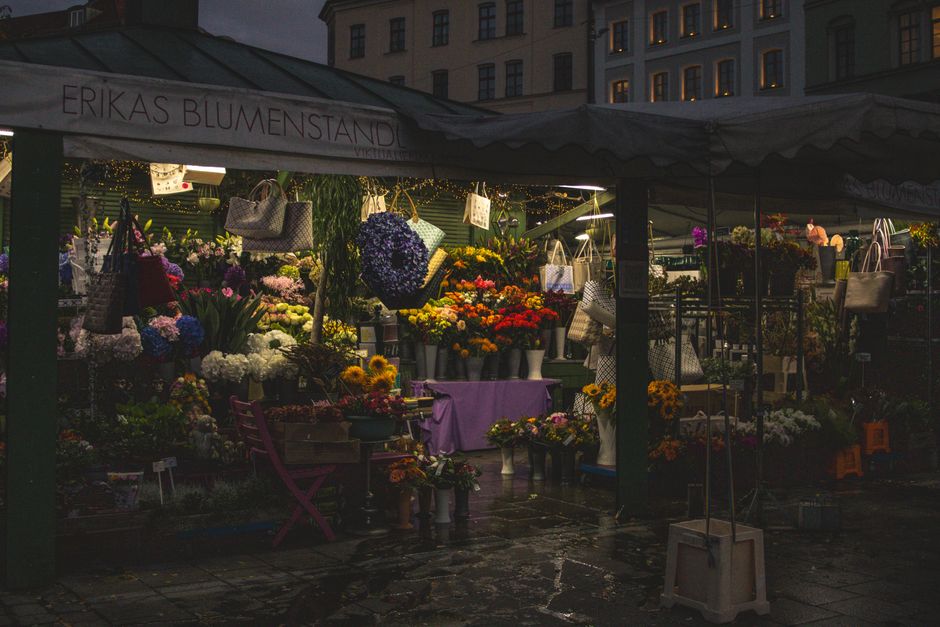 A flower stall under a canopy at dusk, featuring a variety of vibrant fresh floral arrangements and bouquets. The display includes vivid roses, lilies, sunflowers, and hydrangeas in colors ranging fro