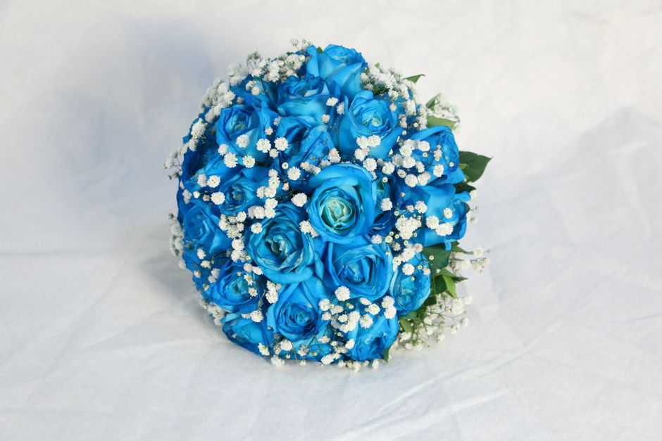 A bouquet of vibrant blue roses with tightly packed petals, accented by small white baby's breath flowers scattered throughout. The bouquet is round and lush, with fresh, slightly glossy green leaves 