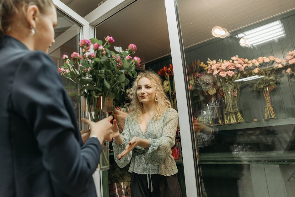 A floral arrangement being handed over by a person wearing a dark blazer to a customer inside a florist shop. The bouquet features pink roses and green foliage, wrapped in beige tissue paper and tied 